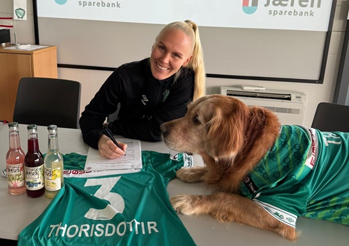 Maria Thorisdottir signing a contract on a table with a green Klepp shirt bearing her surname and number 3, while her dog Theo sits on the table