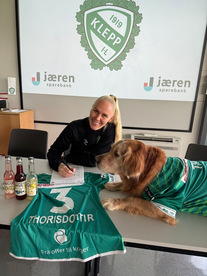 Maria Thorisdottir signing a contract on a table with a green Klepp shirt bearing her surname and number 3, while her dog Theo sits on the table